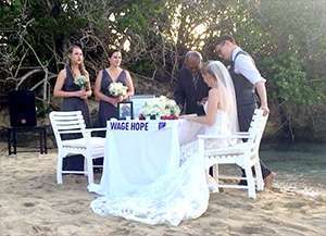 After beachside wedding ceremony, couple sign marriage license at table with Wage Hope banner supporting the cause.