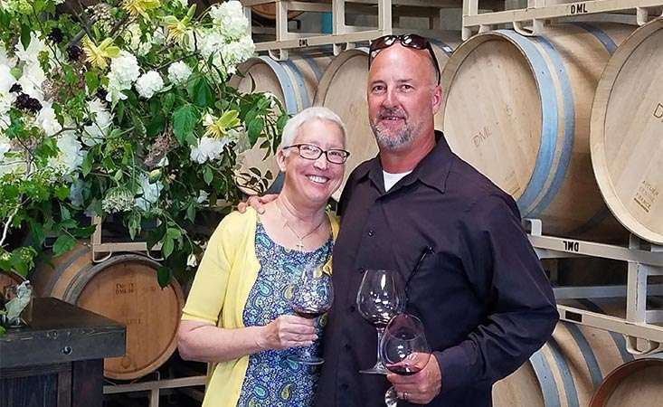 Pancreatic cancer patient and her husband stand before several barrels of their wine company.