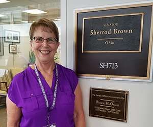 70s Caucasian female standing next to identifying name plate outside a senator's office